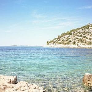 Klares türkisfarbenes Wasser in einer felsigen Bucht mit einem mit Buschwerk bewachsenen Hügel im Hintergrund unter einem strahlend blauen Himmel.