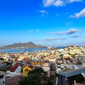 Ein erhöhter Blick auf eine farbenfrohe Küstenstadt mit geschäftigem Hafen und einer Gebirgskette jenseits des Wassers unter blauem Himmel.