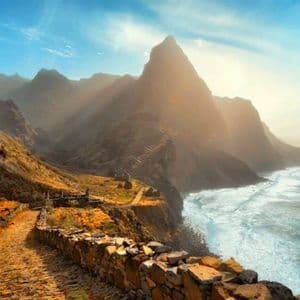 Ein Kopfsteinpflasterweg schlängelt sich entlang einer Klippe mit Meerblick, dahinter erheben sich schroffe Berge unter sonnigem Himmel.