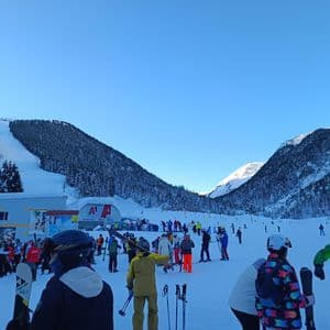 Eine Gruppe von Skifahrern versammelt sich am Fuße eines verschneiten Berghangs mit einer Skiliftstation in der Nähe unter einem klaren blauen Himmel.