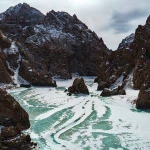 Ein gefrorener Fluss mit türkisblauen Eisflecken schlängelt sich durch eine steile, schneebedeckte Felsenschlucht.