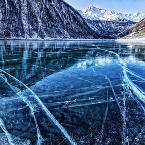 A frozen lake with cracks across its blue ice surface, reflecting the surrounding snow-covered mountains under a clear sky.