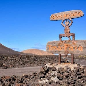 Un cartello di legno per il Parco Nazionale di Timanfaya con il suo simbolo "El Diablo" si trova su rocce vulcaniche accanto a una strada in un paesaggio di campo lavico.