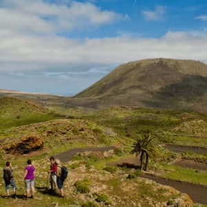 Un viaje en grupo de WeRoad con cuatro personas haciendo senderismo por un sendero a través de un paisaje verde y montañoso, con un gran cono volcánico a lo lejos.