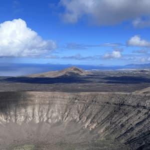 Una vista panorámica de un cráter volcánico en un vasto y oscuro paisaje, con el océano visible a lo lejos bajo un cielo azul y nublado.