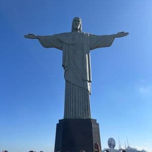 La estatua del Cristo Redentor se eleva sobre una multitud de personas a su base contra un cielo azul claro.