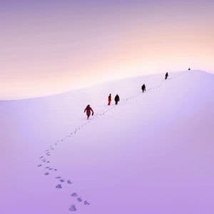 Un viaggio di gruppo WeRoad cammina in fila indiana su un vasto pendio innevato, lasciando impronte sotto un cielo viola e arancione.