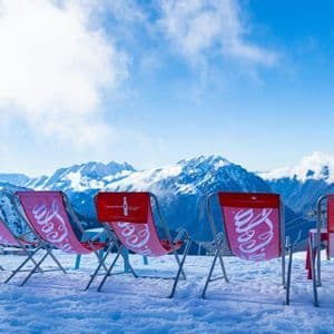 Des chaises longues rouges Coca-Cola, vides dans la neige, font face à une chaîne de montagnes sous un ciel bleu.