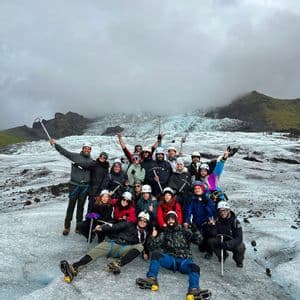 A WeRoad group trip poses for a photo on a glacier, equipped with helmets, crampons, and ice axes under a cloudy sky.