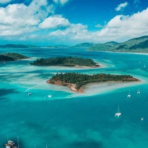 An aerial view of sailboats and catamarans floating in turquoise water surrounding small, forested islands under a blue sky with clouds.