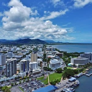 Una vista aérea de una ciudad costera con edificios modernos, un puerto y montañas verdes al fondo bajo un cielo azul parcialmente nublado.