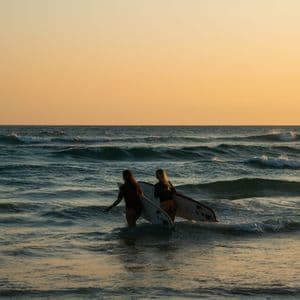 Due donne con tavole da surf camminano tra le onde dell'oceano sotto un caldo cielo al tramonto.