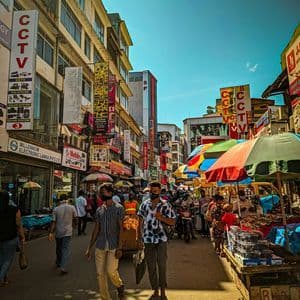 Una strada cittadina affollata con persone che passeggiano davanti a negozi e bancarelle sotto un cielo soleggiato.