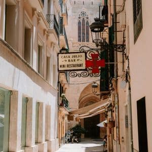 Un estrecho callejón soleado, con edificios históricos y un letrero de restaurante, lleva hacia una iglesia de piedra al fondo.