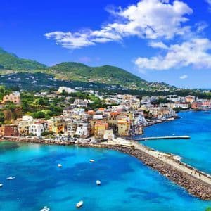 Una vista aerea di una vivace città costiera su una collina, affacciata su una baia blu brillante con un molo in pietra e barche.