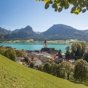 Un villaggio con un campanile si affaccia sulla riva di un lago turchese, con grandi montagne sullo sfondo, visto da una collina erbosa.