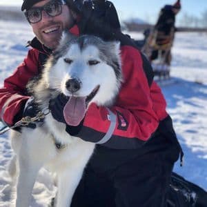 Ein lächelnder Mann mit roter Jacke und Sonnenbrille kniet im Schnee, um einen Husky-Hund mit heraushängender Zunge zu umarmen.
