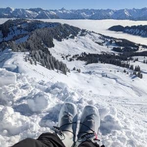 Vue à la première personne de chaussures de ski reposant dans la neige profonde, dominant une vaste chaîne de montagnes enneigée au-dessus d'une mer de nuages.