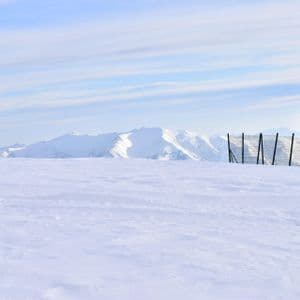 Eine weite, verschneite Landschaft mit einer Holzhütte und einem Zaun rechts sowie schneebedeckten Bergen im Hintergrund.