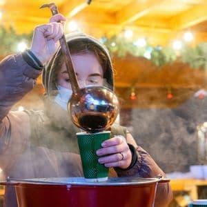 Eine Person in Kapuzenjacke und Gesichtsmaske gießt mit einer großen Kelle ein dampfendes Getränk aus einem großen Topf in eine grüne Tasse.