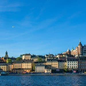 Uno skyline cittadino con edifici storici colorati lungo un lungomare, sotto un cielo azzurro e limpido, con un aereo in volo.