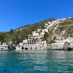Edifici in pietra abbandonati incastonati in un verde pendio, sul bordo di un mare turchese sotto un cielo azzurro e limpido.