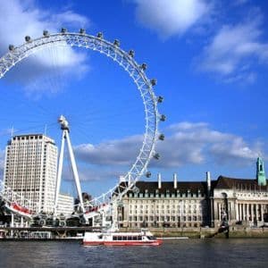 Una grande ruota panoramica bianca sulla riva di un fiume cittadino, con una barca turistica sull'acqua e edifici sullo sfondo sotto un cielo blu.