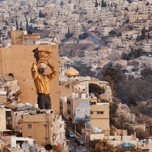Un grande murale di un uomo che porta un capitello di colonna su un edificio domina un paesaggio urbano denso e collinare.