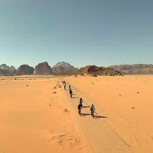 Un gruppo WeRoad in viaggio, pedala in bicicletta in fila su un sentiero sterrato che attraversa un vasto deserto con montagne rocciose in lontananza.