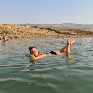 Una donna sorridente galleggia sulla schiena in acqua calma con i piedi sollevati, con una costa rocciosa e arida e colline sullo sfondo.