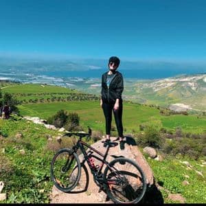 Una donna con l'abbigliamento da ciclismo è in piedi su una roccia accanto alla sua mountain bike, con vista su una valle verde e uno specchio d'acqua.