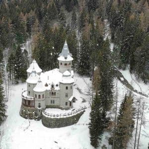 Una vista aerea di un castello di pietra con tetti e torrette innevati immerso in una fitta foresta di pini.