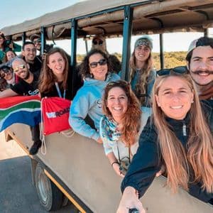 Eine lächelnde WeRoad-Gruppe posiert in einem offenen Safari-Fahrzeug für ein Selfie und zeigt eine südafrikanische Flagge.