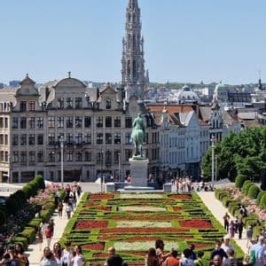 Des gens se promènent dans un jardin formel sur une place de ville, avec des bâtiments historiques et une haute flèche en arrière-plan.