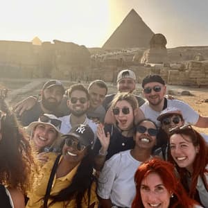 Un voyage de groupe WeRoad prend un selfie souriant avec une pyramide et le Sphinx en arrière-plan au coucher du soleil.