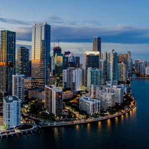Una vista aerea dello skyline di una città moderna con grattacieli illuminati lungo la costa al tramonto.