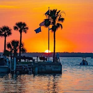 Un tramonto arancione vibrante sull'acqua, con palme e persone in silhouette che osservano da un molo con barche.