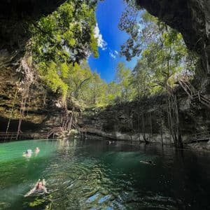 A WeRoad group trip swimming in a large cenote with clear green water, surrounded by rock walls and lush trees under a blue sky.