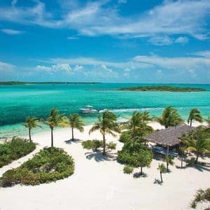 Veduta aerea di una spiaggia tropicale di sabbia bianca con palme mentre un motoscafo naviga sull'oceano turchese sotto un cielo parzialmente nuvoloso.