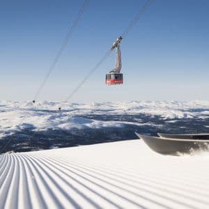 Una funivia viaggia sopra una pista da sci appena battuta, con le punte di uno snowboard in primo piano e montagne innevate sullo sfondo.