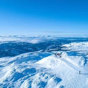 Una vista aerea di un viaggio di gruppo WeRoad su una cima di montagna innevata, che si affaccia su un vasto paesaggio invernale illuminato dal sole sotto un cielo azzurro e limpido.