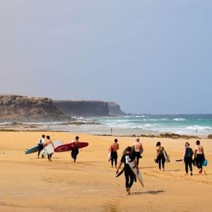 Un groupe WeRoad en combinaisons de surf, planches à la main, marche sur une plage de sable vers l'océan, avec des falaises au loin.