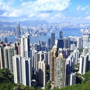 Una veduta dall'alto di un fitto skyline urbano con numerosi grattacieli, adiacente a uno specchio d'acqua, osservata da una rigogliosa collina verde.