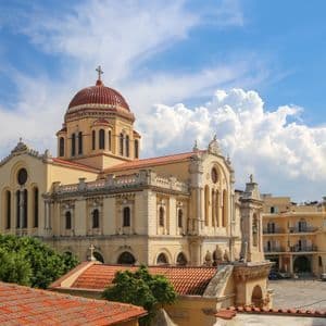 Una gran catedral amarilla con una cúpula roja y un campanario se alza en una plaza de la ciudad bajo un cielo azul nublado.