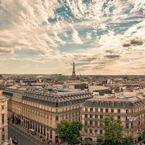Une vue panoramique et plongeante des toits de Paris, avec la Tour Eiffel visible au loin sous un ciel parsemé de nuages.