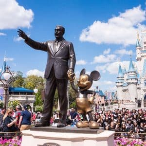 La statue 'Partners' de Walt Disney et Mickey Mouse se dresse devant un château de conte de fées, entourée d'une foule sous un ciel bleu.