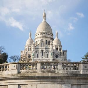 Une grande basilique en pierre blanche avec plusieurs dômes, vue de derrière une balustrade en pierre sous un ciel bleu partiellement nuageux.