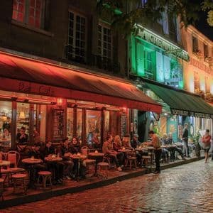 Des gens sont assis aux tables extérieures d'un bar-café, dînant dans une rue pavée la nuit sous des lumières chaudes rouges et vertes.