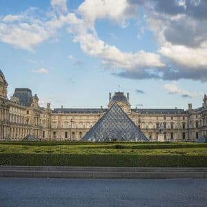 Une grande pyramide de verre se dresse dans la cour d'un bâtiment historique orné, avec une pelouse verte bien entretenue au premier plan sous un ciel nuageux.