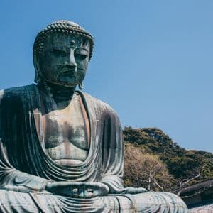 Una grande statua in bronzo di un Buddha seduto all'aperto contro un cielo azzurro limpido, con una collina verde sullo sfondo.